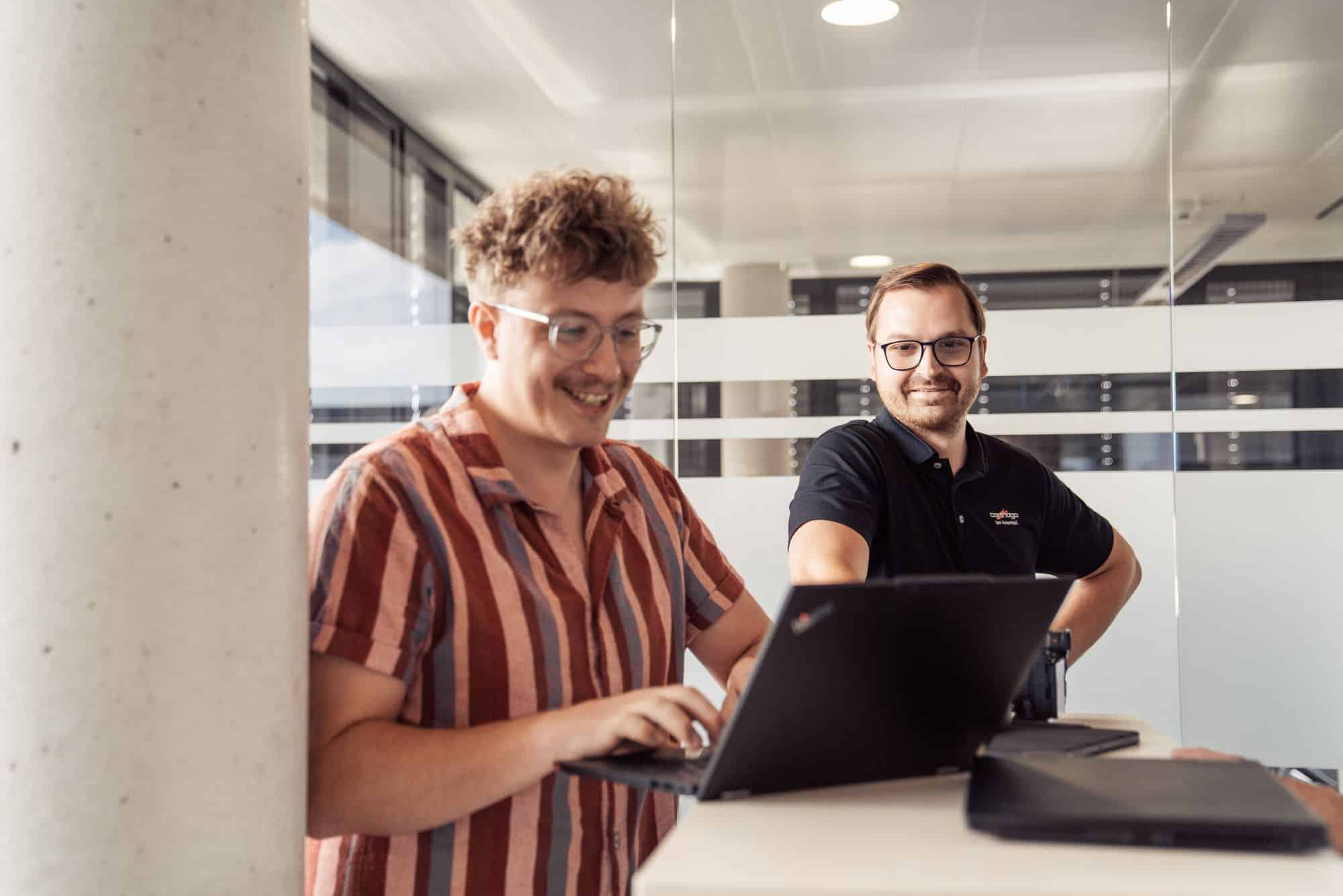Zwei junge Männer arbeiten gut gelaunt gemeinsam an Laptops in einem modernen, hellen Büro mit Glaswänden.