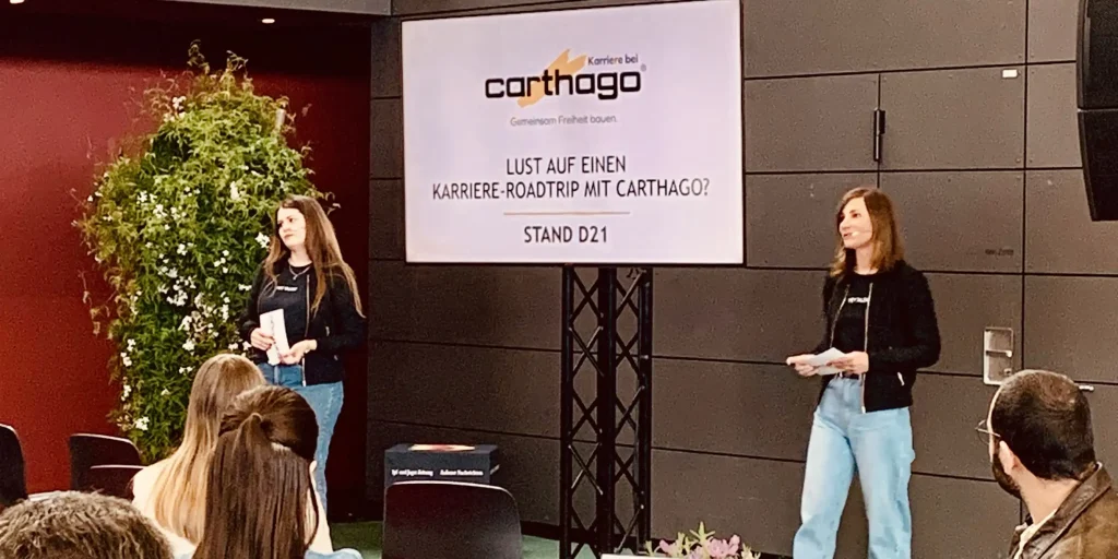 Zwei Carthago-Mitarbeiterinnen präsentieren auf einer Karrieremesse vor Publikum ihren Messestand und laden zum Karriere-Roadtrip ein.