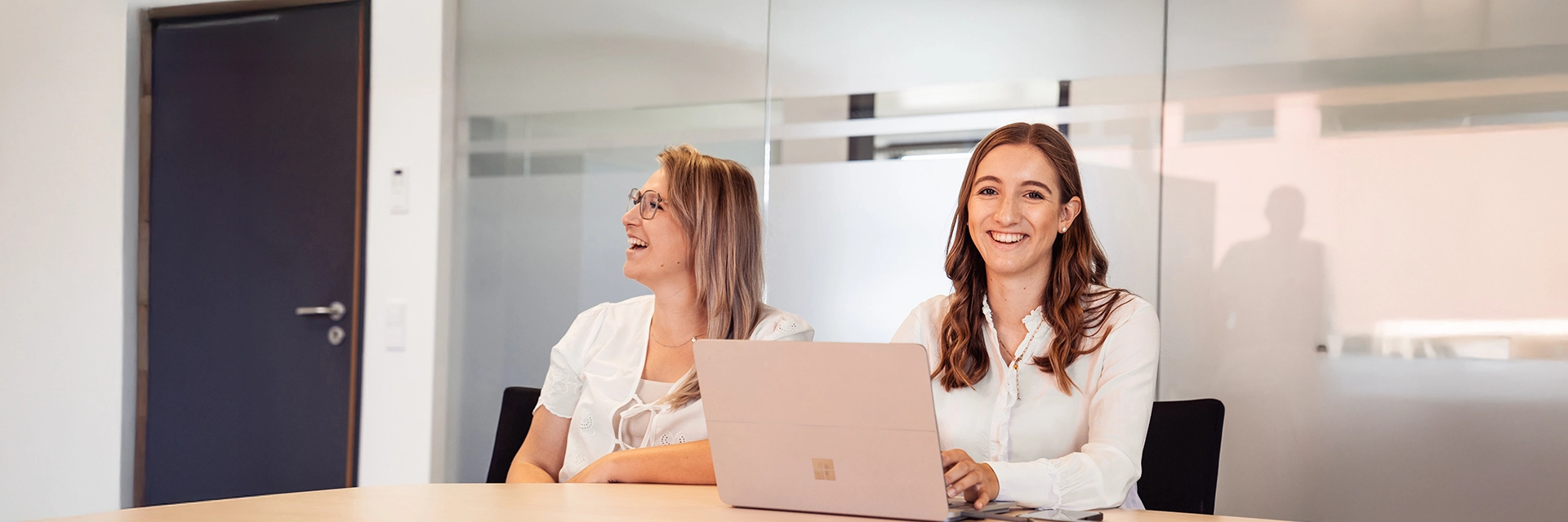 Zwei Frauen sitzen lachend an einem Konferenztisch mit einem Laptop.