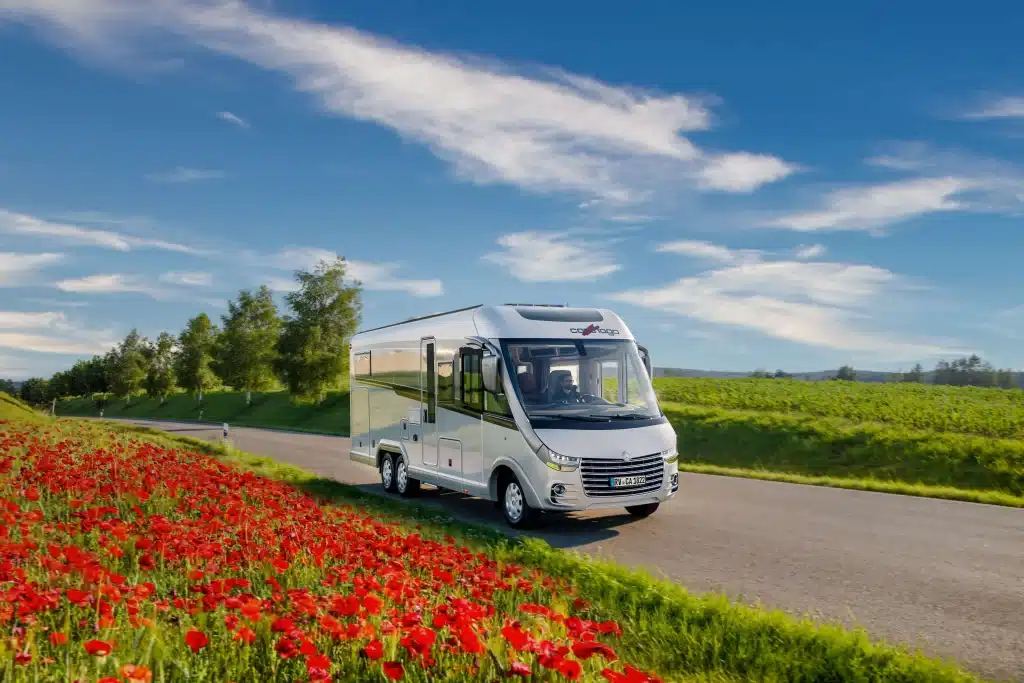 Ein weißes Carthago Reisemobil fährt auf einer sonnigen Landstraße entlang eines roten Mohnblumenfelds, im Hintergrund grüne Wiesen und Bäume.