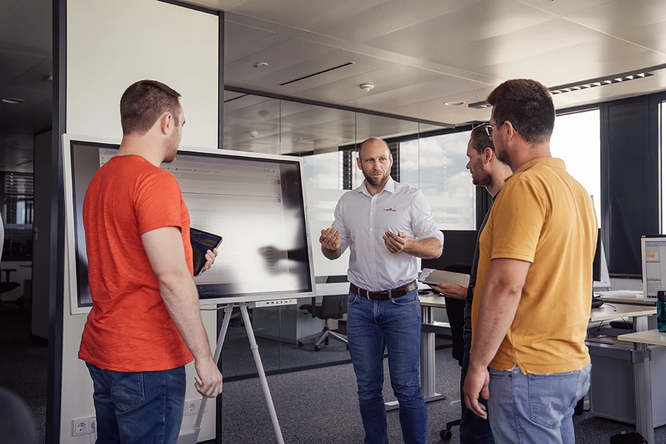 Vier Männer im Gespräch vor einem großen Bildschirm in einem modernen Büro.
