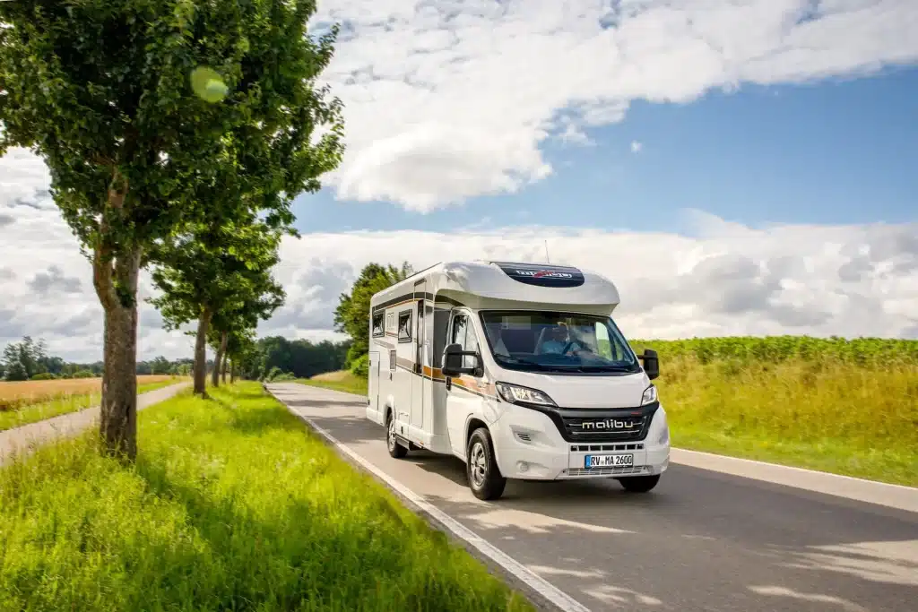 Ein weißes Malibu Reisemobil fährt bei Sonnenschein auf einer schmalen Landstraße durch eine grüne Landschaft mit Bäumen und Feldern.