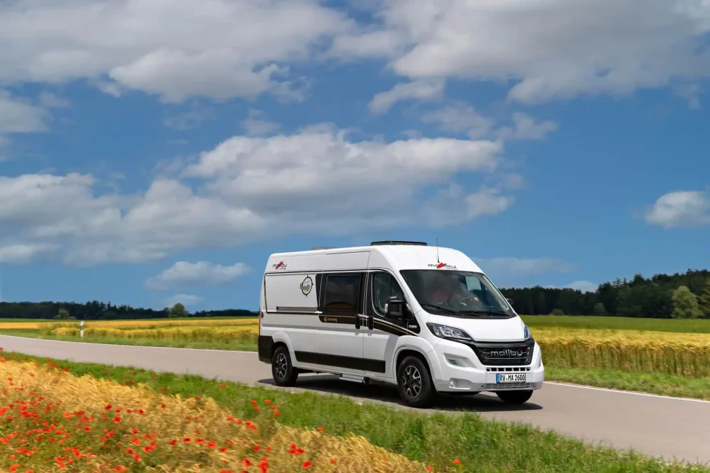 Ein weißer Malibu Van fährt bei Tageslicht durch eine ländliche Gegend mit Getreidefeldern und roten Mohnblumen.