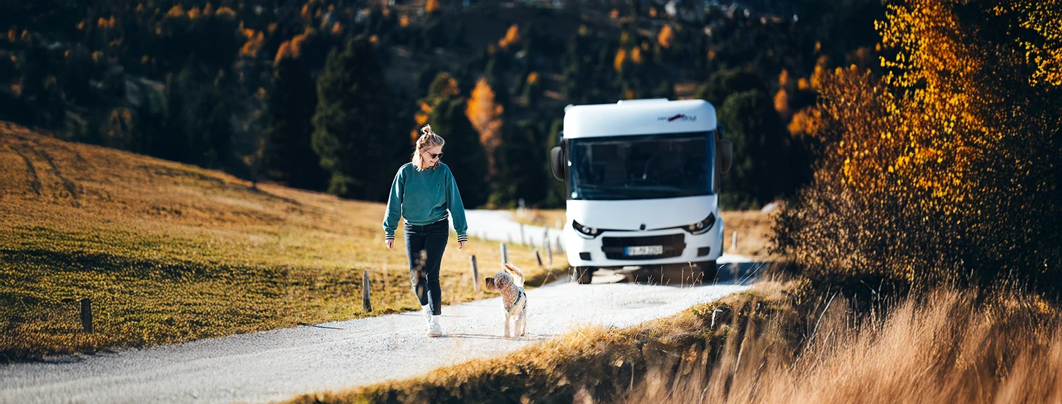Frau geht mit Hund vor einem Wohnmobil durch herbstliche Landschaft spazieren.