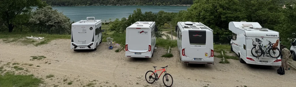 Mehrere Wohnmobile stehen idyllisch auf einem Stellplatz mit Blick auf einen See – bereit für entspannte Tage in der Natur.