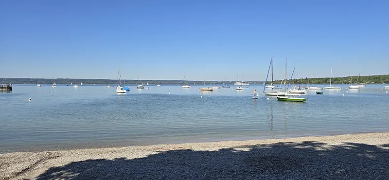 Blick vom Kiesstrand auf einen ruhigen, glitzernden See mit zahlreichen Segelbooten unter strahlend blauem Himmel – ein idyllischer Sommertag in entspannter Atmosphäre.