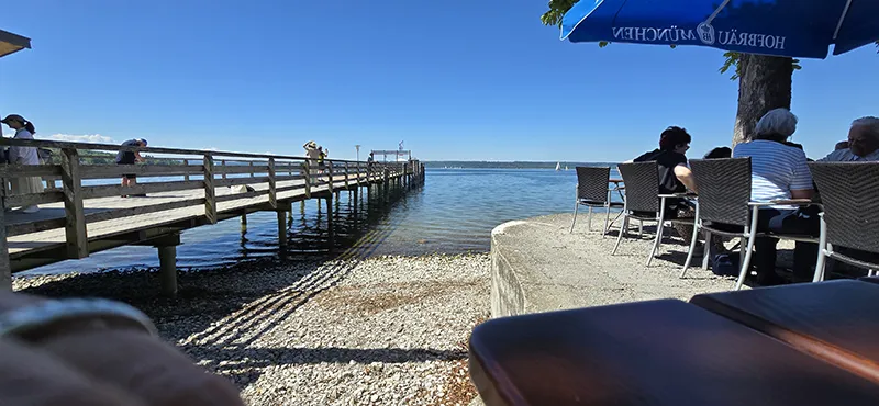 Blick von einem gemütlichen Café mit Sitzplätzen im Schatten eines Sonnenschirms direkt am Seeufer auf einen hölzernen Steg, der in den klaren, blauen See führt – bei strahlendem Sonnenschein und ruhiger Atmosphäre.