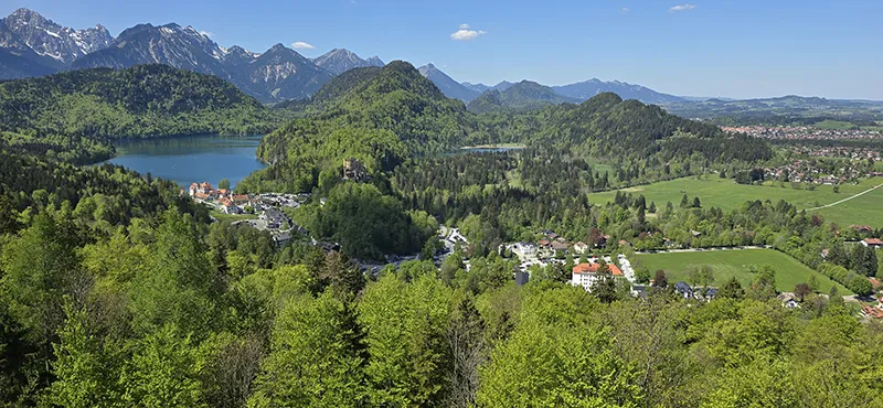 Ein weiter Blick über grüne Hügel, klare Seen und ein kleines Dorf, eingebettet in die beeindruckende Kulisse der Alpen.