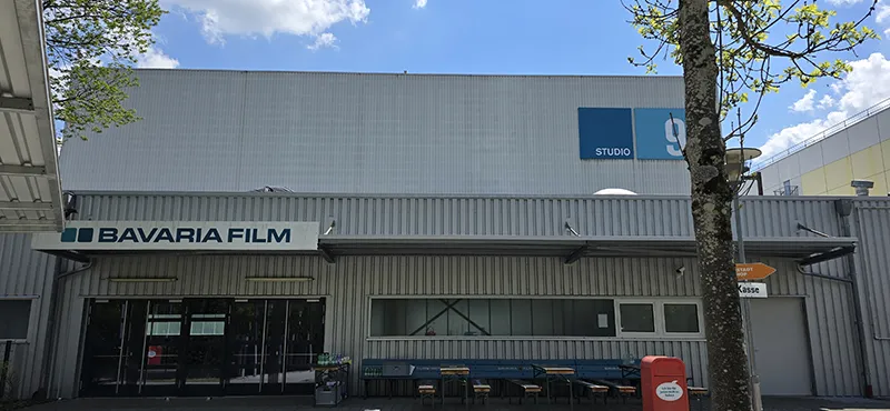 Außenansicht eines Gebäudes der Bavaria Filmstudios mit dem Schriftzug „BAVARIA FILM“ über dem Eingang und einem Hinweisschild zu Studio 9 – davor Sitzbänke, ein Mülleimer und ein Wegweiser zur Kasse, bei sonnigem Wetter.