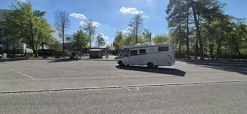 Ein einsamer Stellplatz in der Nachmittagssonne: Ein Carthago-Reisemobil parkt auf einem nahezu leeren Asphaltplatz, umgeben von Bäumen und ruhiger Stadtatmosphäre – bereit für das nächste Abenteuer.