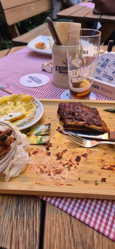 Ein uriger Biergarten-Moment: Auf dem Holztisch mit rot-weiß karierter Tischdecke steht ein deftiges Gericht mit Rippchen, Ofenkartoffel und Zwiebeln – dazu ein halbleeres Erdinger Weißbierglas, ein Steinkrug und Sonnenschein im Hintergrund. Bayrische Gemütlichkeit pur.
