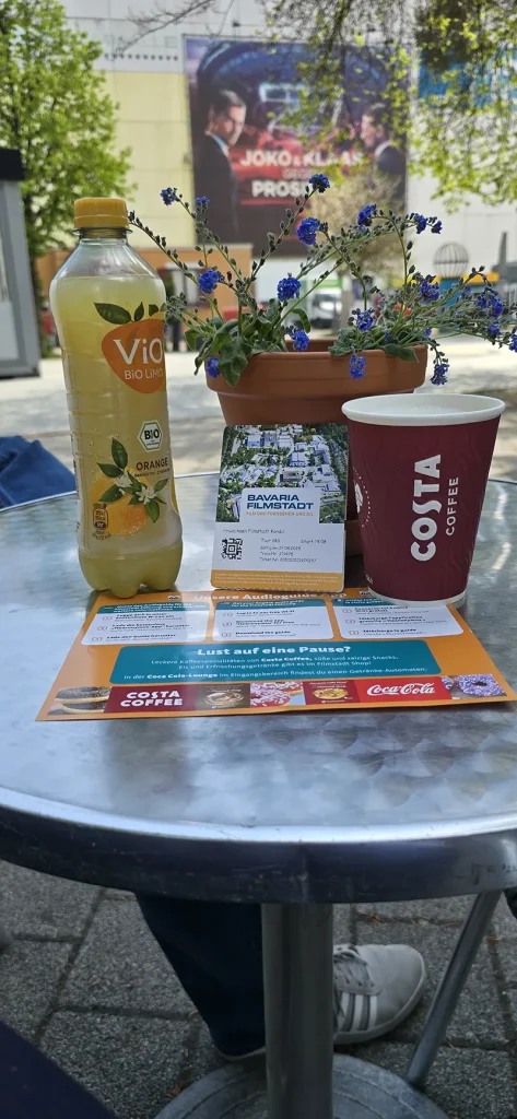 Ein Tisch vor der Bavaria Filmstadt mit einer Flasche ViO Bio Limo, einem Costa Coffee Becher, einem Blumentopf mit blauen Blüten und einem Eintrittsticket zur Filmstadt – im Hintergrund ein Plakat zur TV-Show „Joko & Klaas gegen ProSieben“.