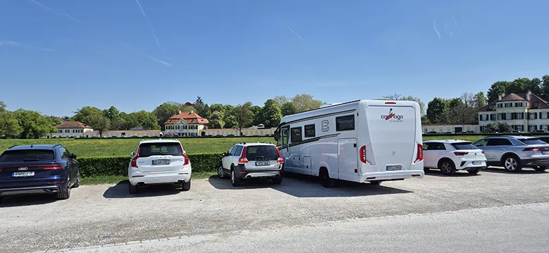 Ein sonniger Parkplatz mit Blick auf gepflegte historische Anwesen und grüne Parkanlagen – mittendrin ein Carthago-Reisemobil, umgeben von modernen PKWs, vor einer Kulisse aus blühender Natur und strahlend blauem Himmel.