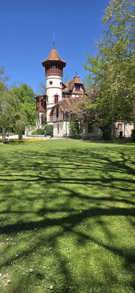 Ein malerisches historisches Gebäude mit Fachwerkelementen und einem runden Turm mit Ziegeldach, umgeben von blühenden Bäumen und einer gepflegten Grünfläche – unter strahlend blauem Himmel und mit klaren Schattenspielen auf dem Rasen.