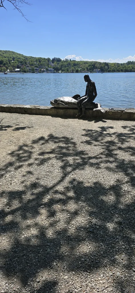 Eine Skulptur einer sitzenden Figur mit einem Fisch am Ufer eines ruhigen Sees, im Hintergrund bewaldete Hügel und vereinzelte Segelboote – unter blauem Himmel mit leichten Wolken und von Blättern beschatteter Uferpromenade.