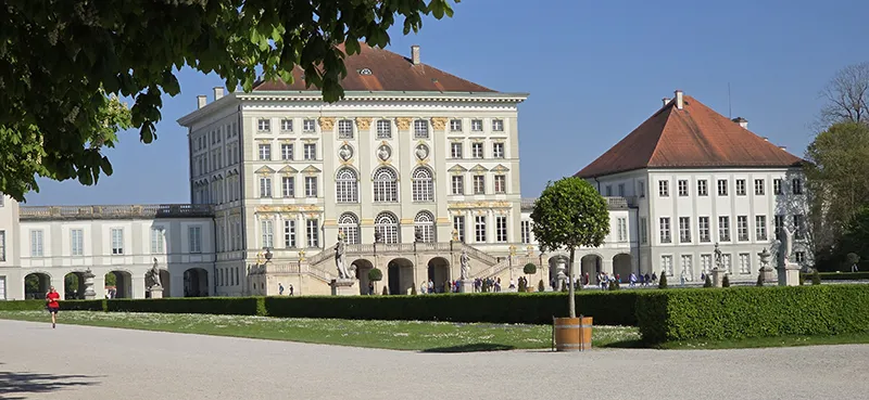 Frontansicht des Schlosses Nymphenburg in München bei sonnigem Wetter, mit klarer Sicht auf die barocke Fassade, gepflegten Gartenanlagen, Spaziergängern und einem Jogger im Vordergrund.