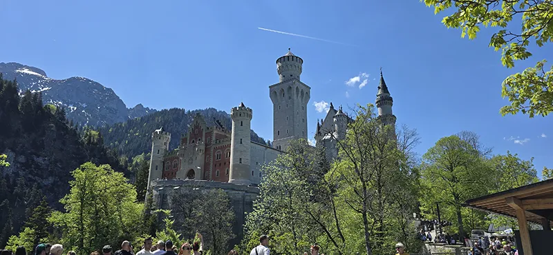Das märchenhafte Schloss Neuschwanstein thront majestätisch über den Baumwipfeln, umgeben von den Alpen und unter strahlend blauem Himmel – im Vordergrund zahlreiche Besucher und grüne Frühlingsbäume.