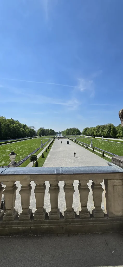 Blick von der Balustrade des Schlosses Nymphenburg auf den symmetrisch angelegten Schlosspark mit breiten Wegen, Springbrunnen, Statuen und gepflegten Grünflächen unter blauem Himmel.