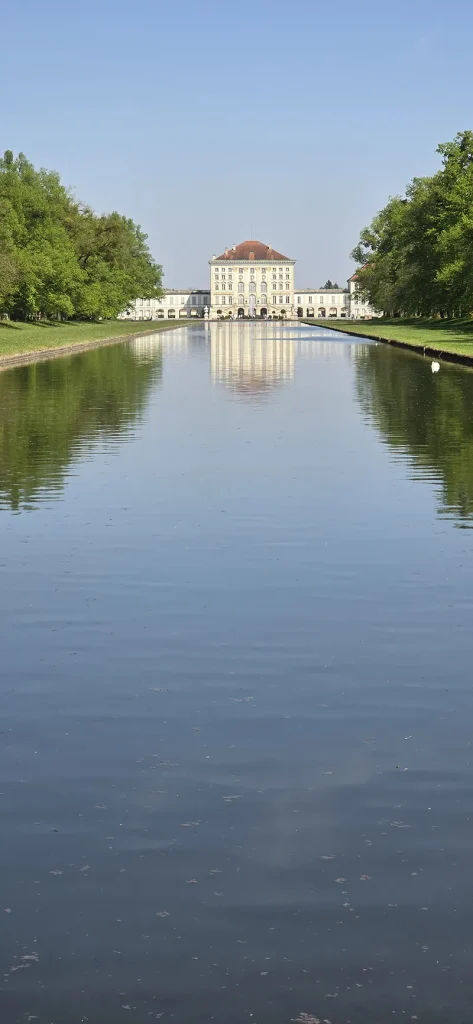 Blick auf das Schloss Nymphenburg in München, das sich malerisch im Wasser des Schlosskanals spiegelt, umgeben von grünen Bäumen und blauem Himmel.