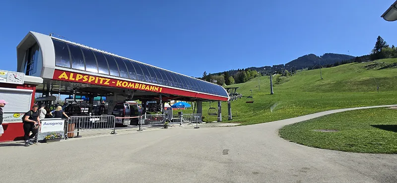 Die Talstation der Alpspitz-Kombibahn 1 bei sonnigem Wetter: Gondeln und Sessellifte fahren in Richtung der grünen Berghänge hinauf, im Hintergrund sind Wanderwege, Wiesen und bewaldete Gipfel zu sehen.