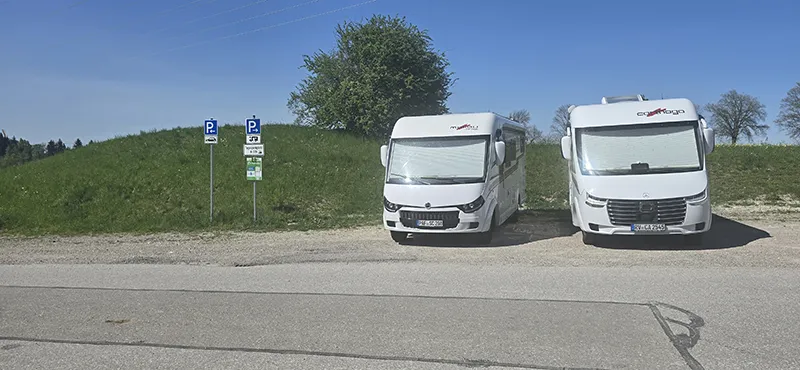 Zwei Wohnmobile – ein Carthago und ein Malibu – stehen nebeneinander auf ausgewiesenen Wohnmobilstellplätzen an einer ruhigen Landstraße vor einem begrünten Hügel, bei strahlend blauem Himmel.