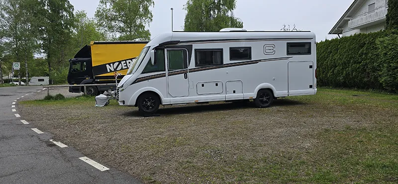 Ein weißes Carthago Wohnmobil steht auf einem befestigten Stellplatz am Rand einer Straße, neben einem gelb-schwarzen LKW und einer gepflegten Hecke – umgeben von Bäumen und typischem Campingplatz-Flair.