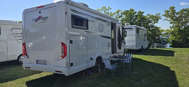 Mehrere Wohnmobile, darunter ein Carthago c-tourer, stehen auf einem grünen, sonnigen Stellplatz am Seeufer. Vor dem Fahrzeug sind Campingstühle und ein Tisch aufgebaut – perfekte Bedingungen für einen entspannten Urlaub in der Natur.