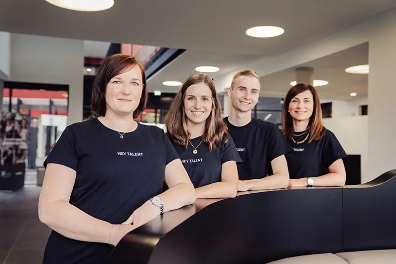 Vier Personen in „HEY TALENT“-Shirts posieren gemeinsam in einem modernen Foyer.