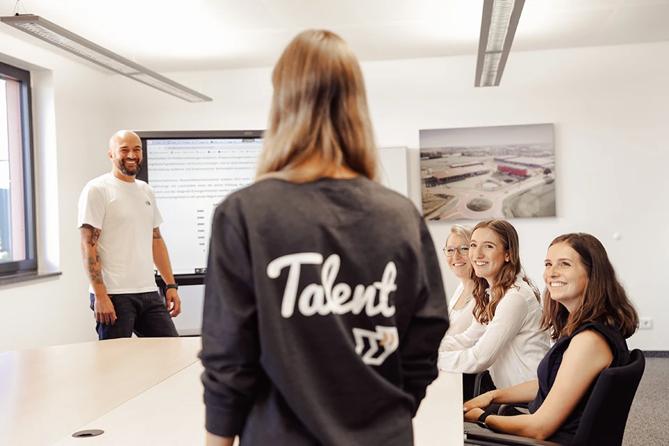 Junge Frau mit „Talent“-Shirt steht vor einer lachenden Gruppe in einem Meetingraum.