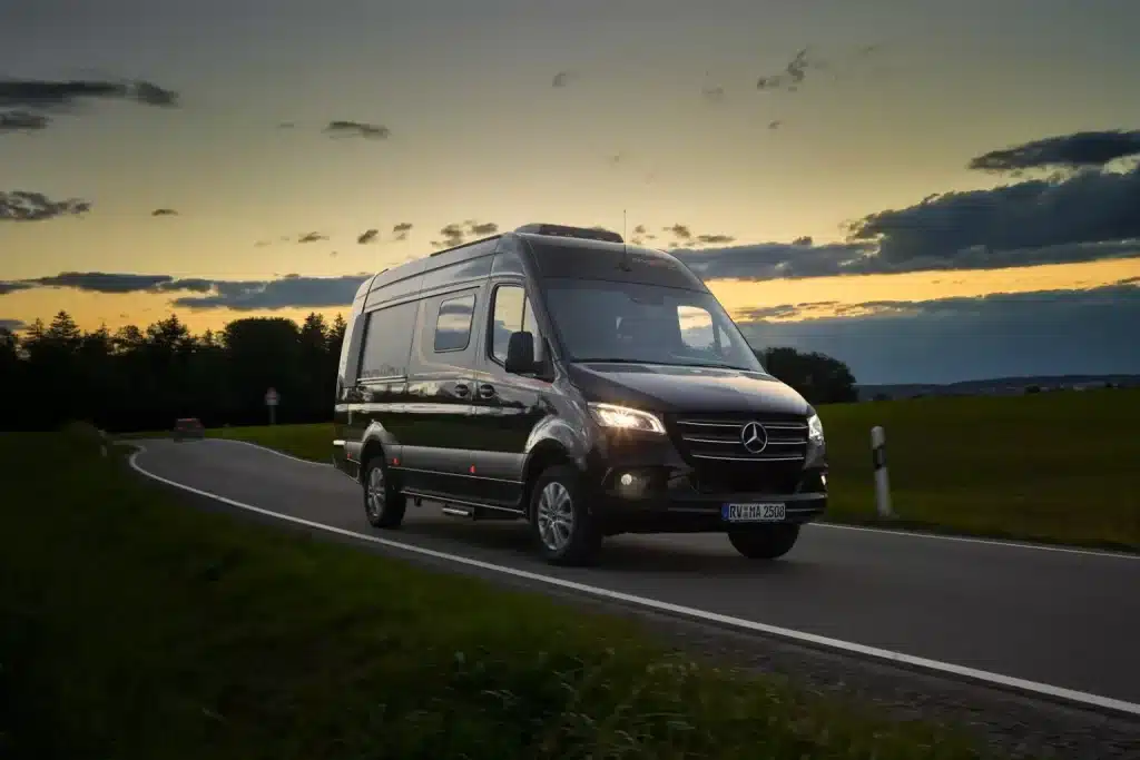 Ein schwarzer Malibu Van auf Mercedes-Benz-Basis fährt über eine kurvige Landstraße im Abendlicht.