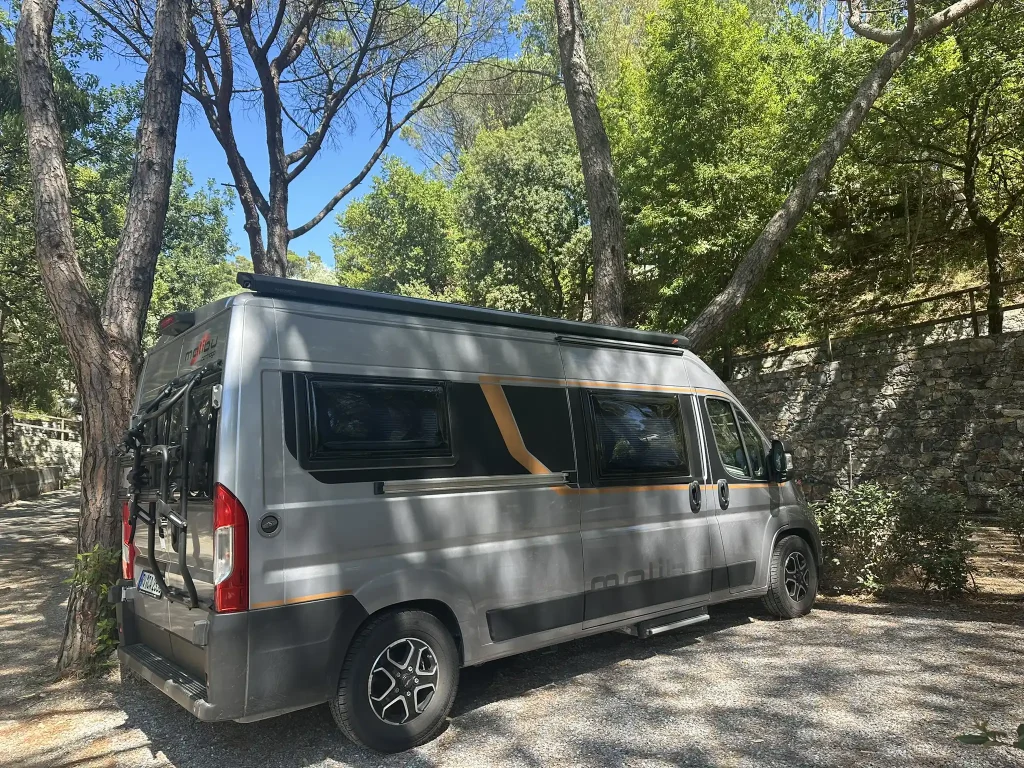 Malibu Van auf einem Stellplatz umgeben von Bäumen