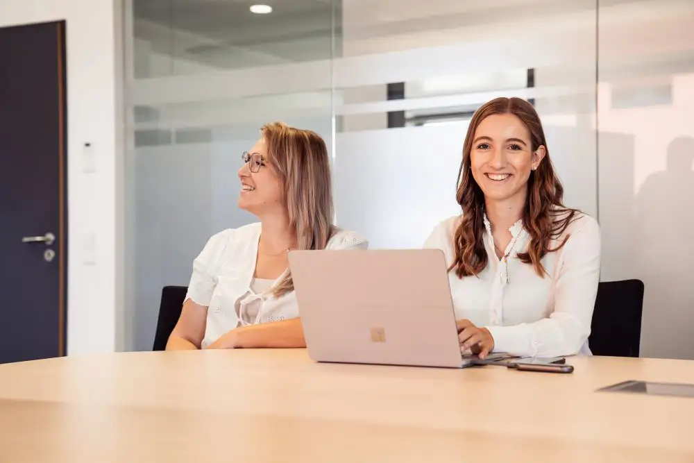 Eine Mitarbeiterin am Laptop und eine weitere Mitarbeiterin neben ihr am Tisch, die in den Bildschirm schaut