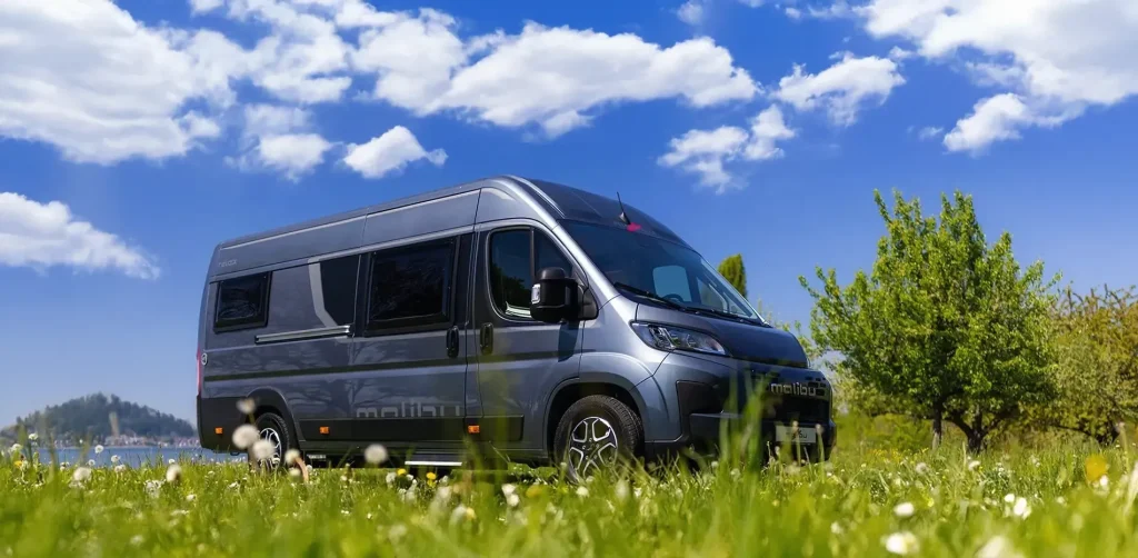 Der neue Premium Van steht auf einer grünen Wiese mit Blumen, im Hintergrund Bäume, ein See und ein blauer Himmel mit weißen Wolken.